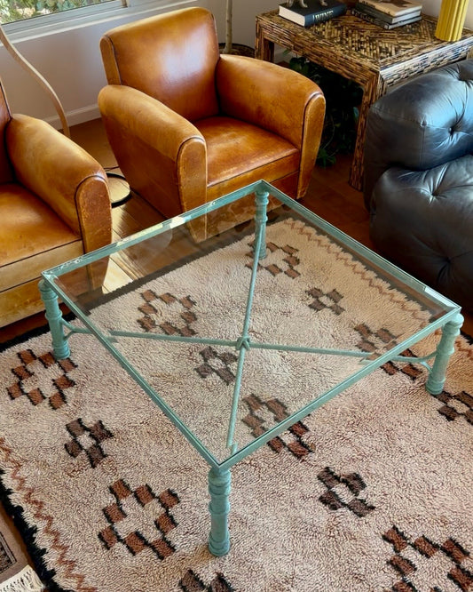 Green verdigris metal coffee table with beveled glass top, in the manner of Diego Giacometti, styled on a patterned rug beside vintage leather club chairs