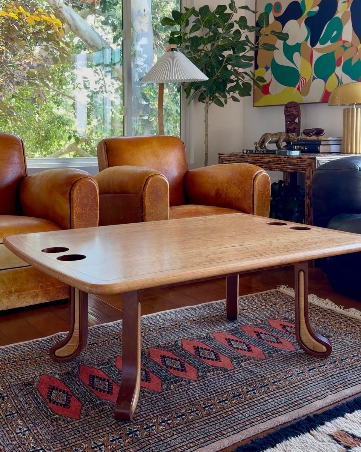 Two-tone sculpted studio craft coffee table with built-in cup holders, styled on a patterned rug in a sunlit living room
