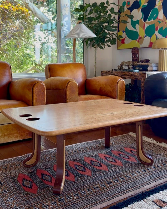 Two-tone sculpted studio craft coffee table with built-in cup holders, styled on a patterned rug in a sunlit living room